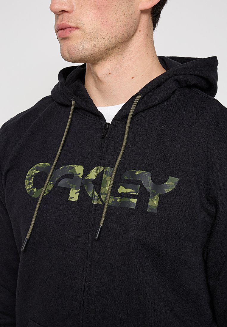 Felpa nera con zip e logo "Oakley" in camouflage, dotata di cappuccio con cordoncino e tessuto testurizzato. Cordoncini regolabili in verde oliva.
