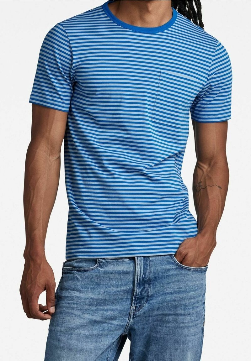 Homme portant un t-shirt à manches courtes à rayures horizontales bleues et blanches avec une poche poitrine et un jean en denim bleu clair, main dans la poche.