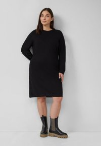 Frau, die ein langes schwarzes Strickkleid und schwarze Stiefeletten mit beigen Sohlen trägt, steht vor einer schlichten hellgrauen Wand.