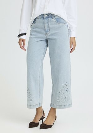 Woman wearing light blue wide-leg cropped jeans with floral embroidery near hems, paired with white blouse and black heeled slingback shoes.