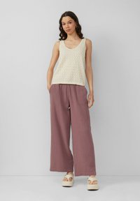 Licht beige gebreide tanktop met V-hals, gecombineerd met brede mauve broekspijpen. Model in platform sandalen, staand tegen een grijze achtergrond.
