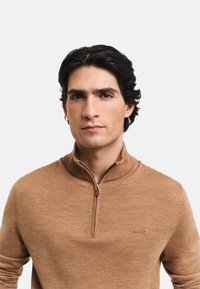 Suéter marrón con cuello de cremallera, hecho de una tela suave, con una textura sutil, diseño ajustado y un pequeño logo en el pecho.