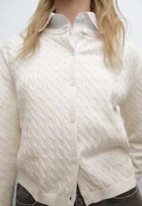 Weißer Strick-Cardigan mit einem strukturierten Muster, mit einer Knopfleiste und einem gerippten Saum, über einem Hemd mit Kragen getragen.