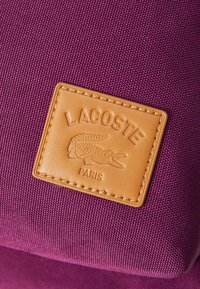 Sac en textile violet avec un patch en cuir beige portant un logo de crocodile et "LACOSTE PARIS" embossé. Matériau texturé, forme carrée.