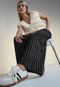 Gilet lavorato a maglia color crema con scollo a V, chiusura con bottoni e lavorazione a rete aperta; pantaloni a gamba larga con righe nere; sneakers bianche con dettagli neri e suole marroni.