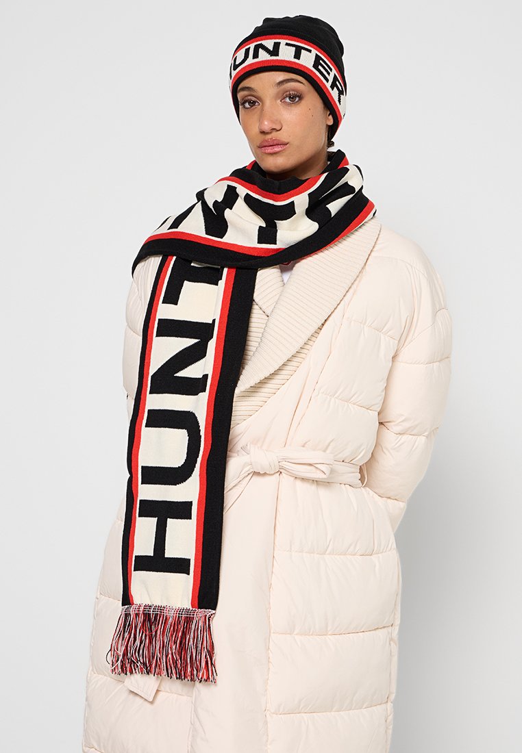 Czarny i kremowy dziany szalik z widocznym napisem "HUNTER" w pogrubionej czcionce, z czerwonymi akcentami i frędzlami, noszony na kremowej kurtce puchowej.
