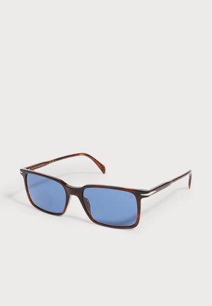 Lunettes de soleil rectangulaires en écaille avec des branches fines et des verres teintés bleus posées sur une surface blanche.
