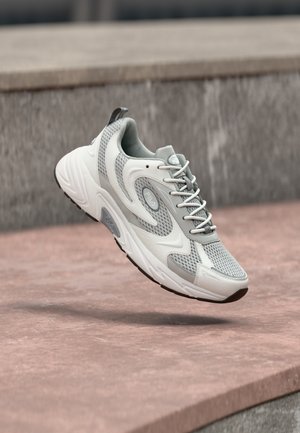 Bela in siva športna superga z mrežastimi in usnjenimi detajli, v zraku nad kamnito površino, prikaz strani in zgornjega pogleda.