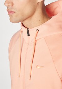 Lys fersken zip-up hettegenser laget av mykt stoff. Har justerbare snorer, svart glidelås og en gull Nike-logo på brystet.