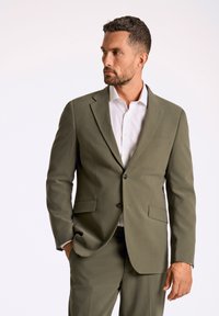 Olijfgroen pak gemaakt van gestructureerde stof; enkel breasted blazer met twee zakken en een knopen sluiting, gecombineerd met een wit shirt.