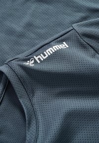 Mørkegråt åndbart sportsstof med små perforeringer og hvidt "hummel" logo printet på overfladen nær en søm.