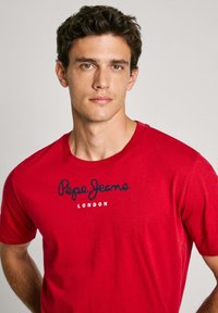 Rød bomulds T-shirt med rund halsudskæring og korte ærmer, med "Pepe Jeans London" trykt i sort skrift på forsiden.