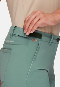 Türkise bukser med en glat tekstur, der har et bælte med Velcro-lukning og en sidelomme med lynlås. Logo nær taljen.