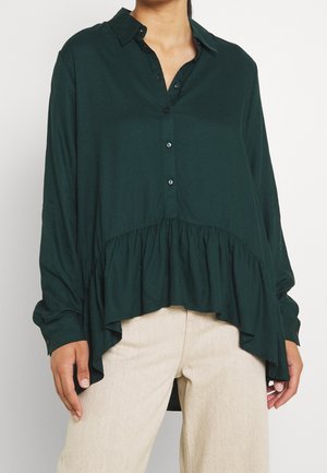 Donna che indossa una camicia botton-up verde scuro con maniche lunghe e orlo arricciato a lunghezze variabili, abbinata a pantaloni beige chiaro.