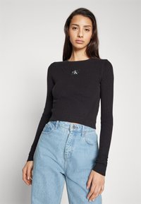 Sort ribbet langærmet top med rund hals og et lille sort logo patch. Sammen med lyseblå denim jeans.