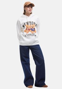 Sudadera blanca con un gráfico de dos osos, combinada con jeans de pierna ancha de color azul oscuro y una gorra color beige. El atuendo tiene un ajuste casual y relajado.