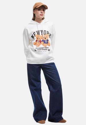 Un hoodie blanc avec un graphisme de deux ours, associé à un jean large bleu foncé et une casquette beige. La tenue a une coupe décontractée et confortable.