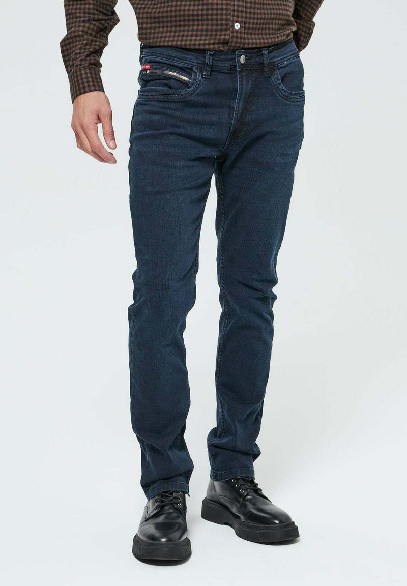 Jean en denim bleu foncé avec une coupe slim, dotés de poches avant, d'une poche zippée sur la cuisse et d'une fermeture à bouton. Portés avec des chaussures noires.