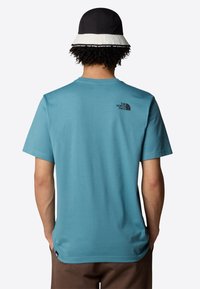 Camiseta de algodón turquesa de manga corta y cuello redondo. Presenta un logo negro en la parte trasera y se combina con un sombrero tipo bucket en blanco y negro.