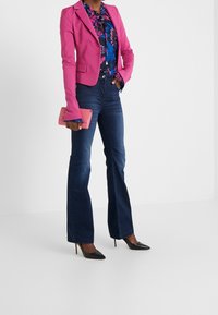 Žena v růžovém saku a květované blůze s tmavými rozšířenými džíny a černými vysokými podpatky, držící růžovou psaníčko.