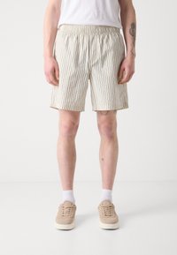 Pantalones cortos a rayas beige con cintura elástica, con rayas oscuras verticales. Combinados con zapatillas beige y calcetines blancos.