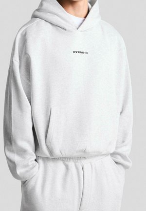 Persoon draagt een lichtgrijze oversized hoodie en bijpassende joggingbroek met het logo "OVRNARM" op de borst van de hoodie, handen in de zakken.