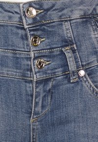 Vaqueros de mezclilla azul de cintura alta con tres botones metálicos, costuras dobles y detalles en los bolsillos frontales. Textura suave y acabado desgastado.