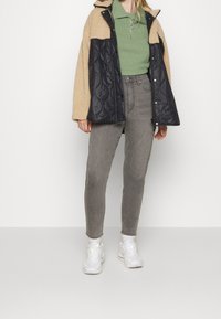 Chaqueta de forro polar acolchada en negro y beige con cremallera frontal, combinada con una camiseta polo verde y vaqueros ajustados grises. Zapatillas blancas completan el conjunto.