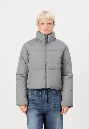 Puffy grå jakke med høy krage, frontglidelås og elastisk kant. Har sydd seksjoner og vide ermer. Kombinert med blå denim jeans.