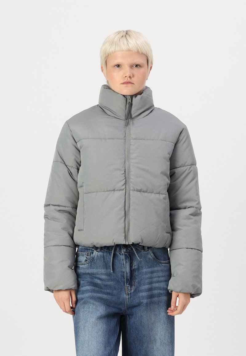 Giacca grigia gonfiata con colletto alto, zip anteriore e orlo elastico. Presenta sezioni cucite e maniche larghe. Abbinata a jeans in denim blu.