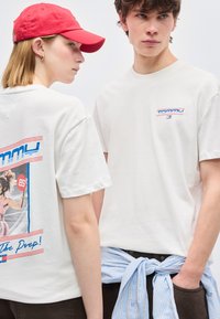 Δύο νεαροί ενήλικες φορούν λευκά t-shirts Tommy Jeans, ο ένας με κόκκινο καπέλο και γραφική εκτύπωση στην πλάτη, ο άλλος με ριγέ πουκάμισο δεμένο γύρω από τη μέση.