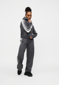 Σετ γκρι φούτερ με κουκούλα και παντελόνι φόρμας με λευκές ρίγες στο πλάι, απαλής υφής και άνετης εφαρμογής. Το μοντέλο φοράει μαύρα παπούτσια.