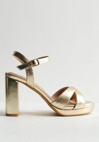 Le sandali platform metallici dorati presentano un design con una larga fascia, chiusura con fibbia alla caviglia e un alto tacco a blocco per maggiore altezza e stabilità.