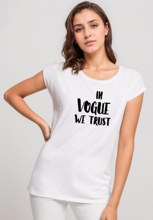 Young woman posing with hand on hip wearing a white t-shirt with black text "IN VOGUE WE TRUST" on the front.