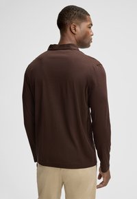 Braun, langärmliges Poloshirt mit Kragen, aus weichem Stoff gefertigt. Hat einen legeren Schnitt und dezente Stickdetails am Saum.