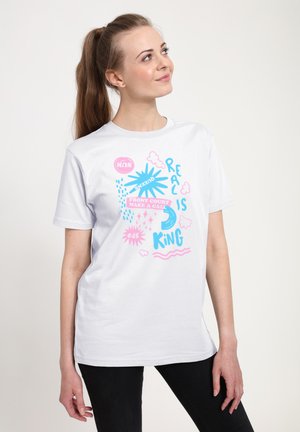 Kvinde med brunt hestehale iført hvid T-shirt med blå og pink grafisk tekst, inklusive "Real is King" og "Front court make a call."
