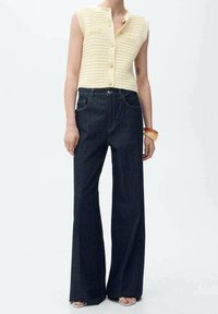 Dzianinowy top bez rękawów w jasnżółtym kolorze z zapięciem na guziki, zestawiony z ciemnoniebieskimi, rozszerzanymi dżinsami z wysokim stanem. Gładka tekstura, minimalne detale.