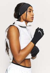 Sorte Nike handsker med et reflekterende logo, båret med en hvid puffy vest. Har struktureret stof og et tætsiddende design. Korte hår, profilbillede.