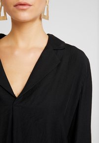 Chemise noire à col avec un décolleté en V profond et une texture lisse, complétée par de longues boucles d'oreilles géométriques en or.