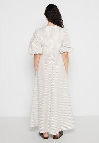 Femme aux longs cheveux bouclés portant une robe blanche longue jusqu'aux chevilles avec de fines rayures verticales noires et des manches bouffantes, debout pieds nus.