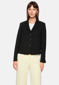 Schwarzer taillierter Blazer mit Reverskragen, drei Knöpfen, Seitentaschen und kontrastierenden Nähten, getragen über einem weißen Oberteil und cremefarbenen Hosen.