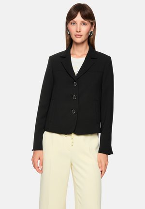 Schwarzer taillierter Blazer mit Reverskragen, drei Knöpfen, Seitentaschen und kontrastierenden Nähten, getragen über einem weißen Oberteil und cremefarbenen Hosen.