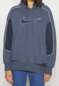 Navy blue pullover hoodie with white accents, hood, and front pouch pocket. Features a large white Nike logo on the chest and ribbed cuffs.