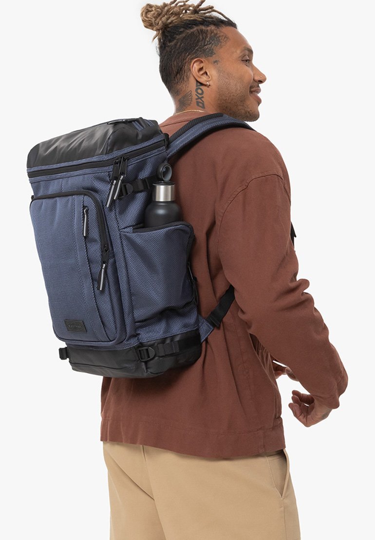 Sac à dos marine avec un tissu texturé et des accents noirs, doté de plusieurs compartiments zippés et d'une poche latérale pour une bouteille d'eau.