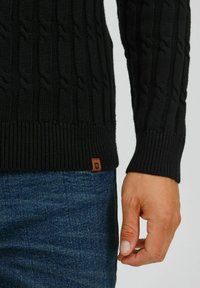 Pull noir en tricot avec un motif en relief, poignets et ourlet côtelés, avec un petit label marron près du bas. Porté avec un jean bleu.