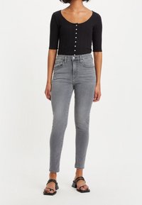 Top negro de manga larga con relieve y cierre de botones, combinado con jeans ajustados grises de cintura alta y sandalias negras con tiras.
