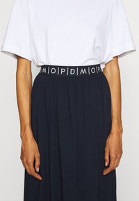 Jupe navy avec une large ceinture élastique arborant un texte logo blanc. Le tissu semble doux avec un design fluide et plissé. T-shirt blanc visible.