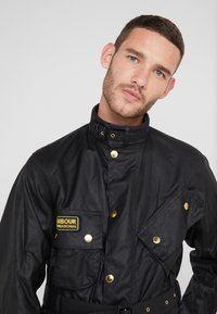 Black jacket made of textured fabric, featuring gold hardware, multiple pockets, a high collar, and a belted waist design.