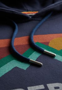 Marineblauwe hoodie van textuurstof, met dikke trekkoorden in donkerblauw en metalen uiteinden. Multikleurige geometrische patronen eronder.