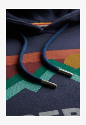 Marineblauwe hoodie van textuurstof, met dikke trekkoorden in donkerblauw en metalen uiteinden. Multikleurige geometrische patronen eronder.